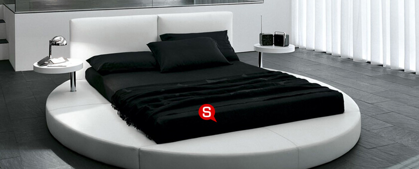 Sypialnia w stylu futurystycznym z czarno-białym łóżkiem na okrągłej podstawie. Po jego obu stronach znajdują się stoliki kawowe na metalowych podstawach.