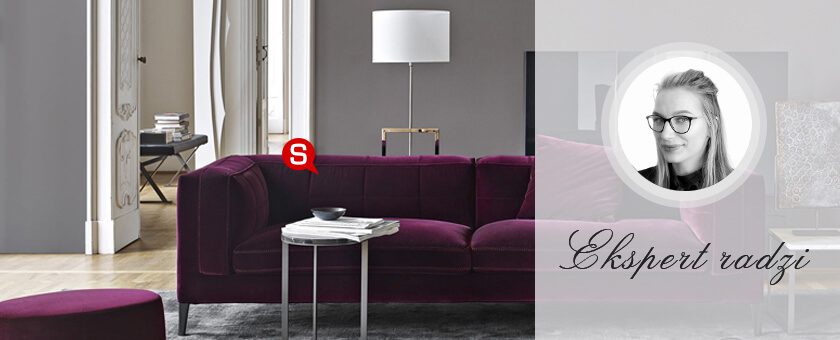 Klasyczny salon z szarymi ścianami oraz kanapą i pufą w kolorze purpury. Całości aranżacji dopełnia biała lampa stojąca ze złotymi elementami oraz subtelny stolik kawowy.
