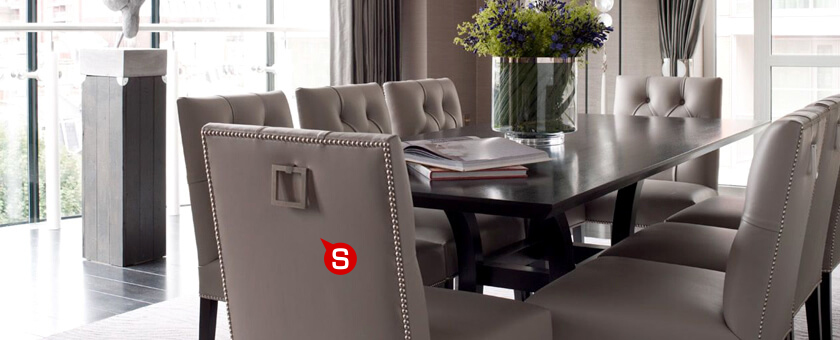 Jadalnia w stylu klasycznym z dużym drewnianym stołem, wokół którego znajdują się eleganckie, siwe, tapicerowane krzesła z elementami ozdobnymi. Całości aranżacji dopełniają akcenty florystyczne.