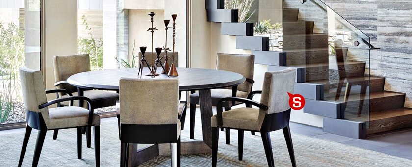 Jadalnia w stylu modernistycznym z dużym okrągłym stołem oraz tapicerowanymi krzesłami. Na stole widać nowoczesne świeczniki. W głębi pomieszczenia znajdują się schody ze szklanymi elementami. Całość utrzymana jest w odcieniach brązów.