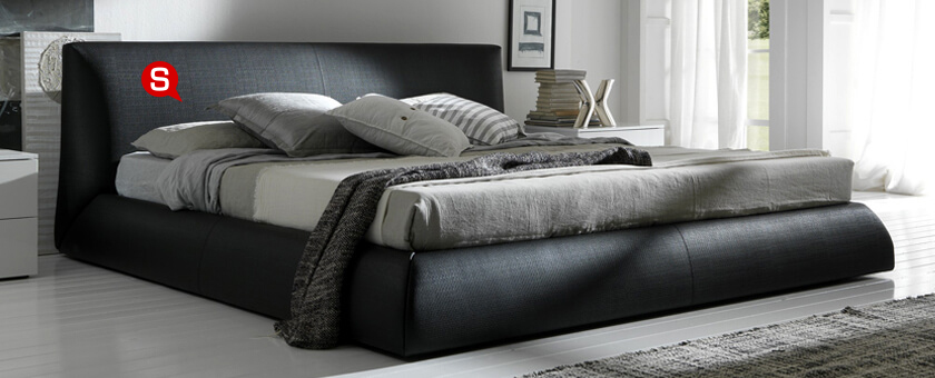 Duże, czarne łóżko kontynentalne w przestrzennej sypialni. Na szafce nocnej znajduje się srebrna dekoracja. Całość utrzymana jest w minimalistycznych kolorach, takich jak czerń, szarość i biel.